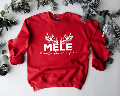 A red sweater with the text "MELE KOLIKINAS" printed on it, surrounded by pine branches and greenery.