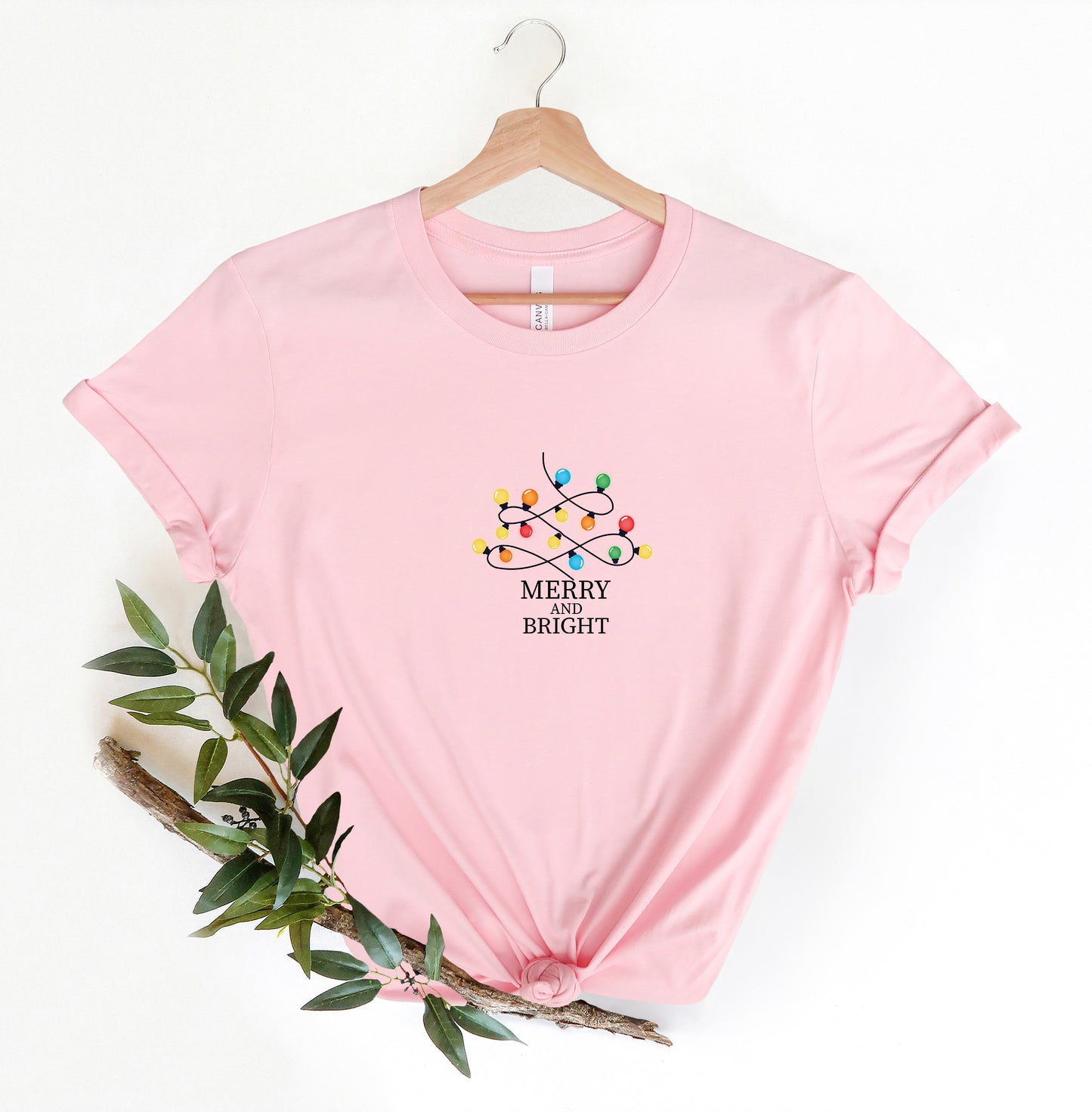 A pink t-shirt with a graphic design of colorful Christmas lights and the text "MERRY BRIIGHT" printed on it, hanging on a wooden hanger.
