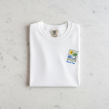 A white t-shirt with a small graphic design and text printed on it, laid out on a marble surface.