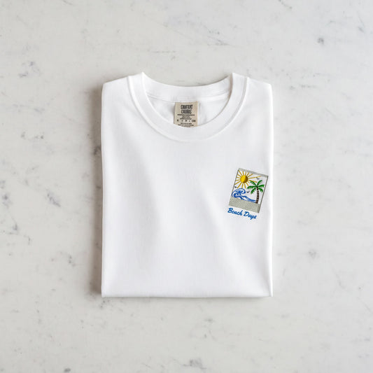 A white t-shirt with a small graphic design and text printed on it, laid out on a marble surface.