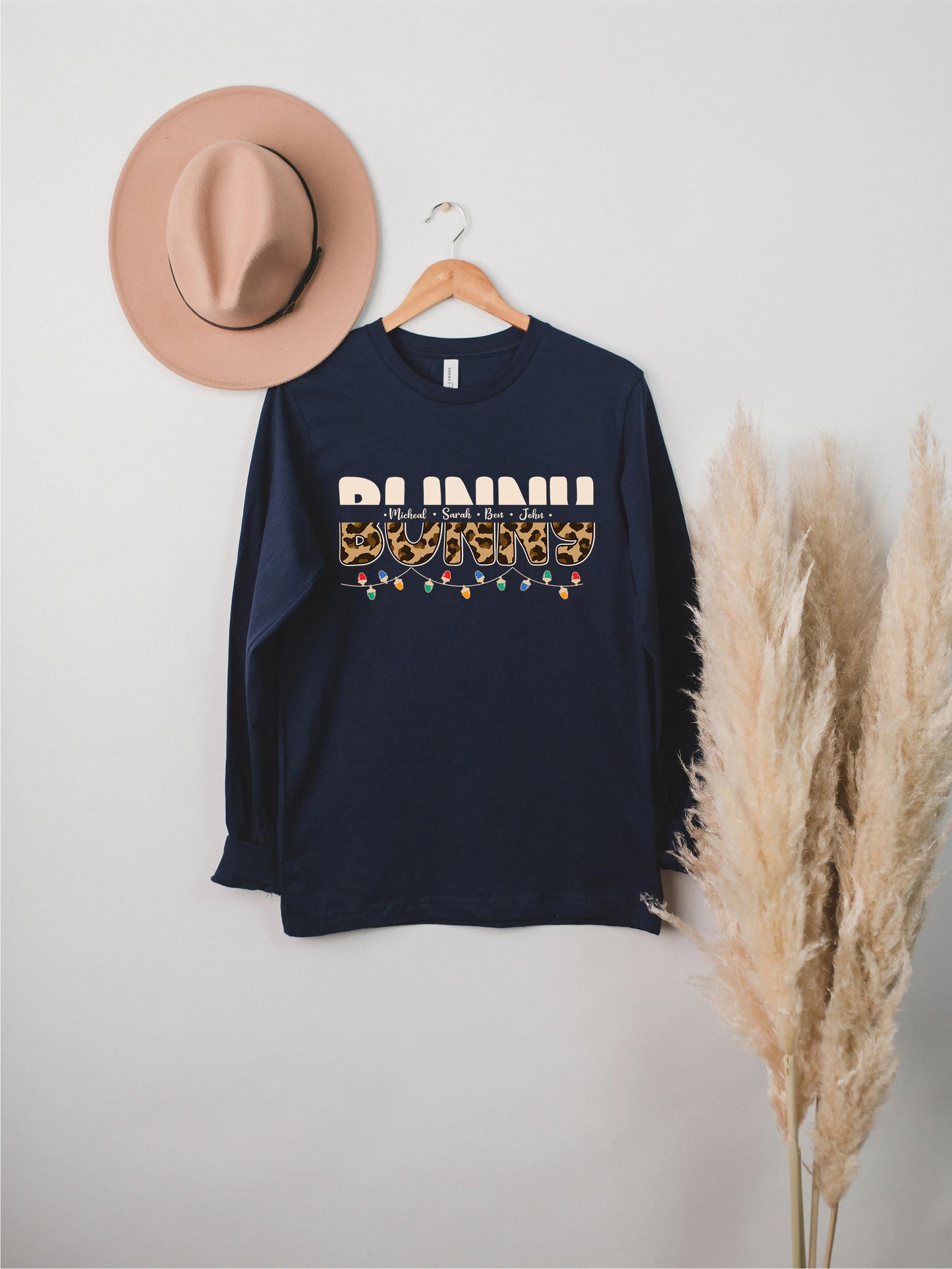 A navy blue long-sleeved shirt with the word "BUNNI" printed on it, hanging on a wooden hanger. Next to the shirt is a beige hat, and there are some potted plants in the background.