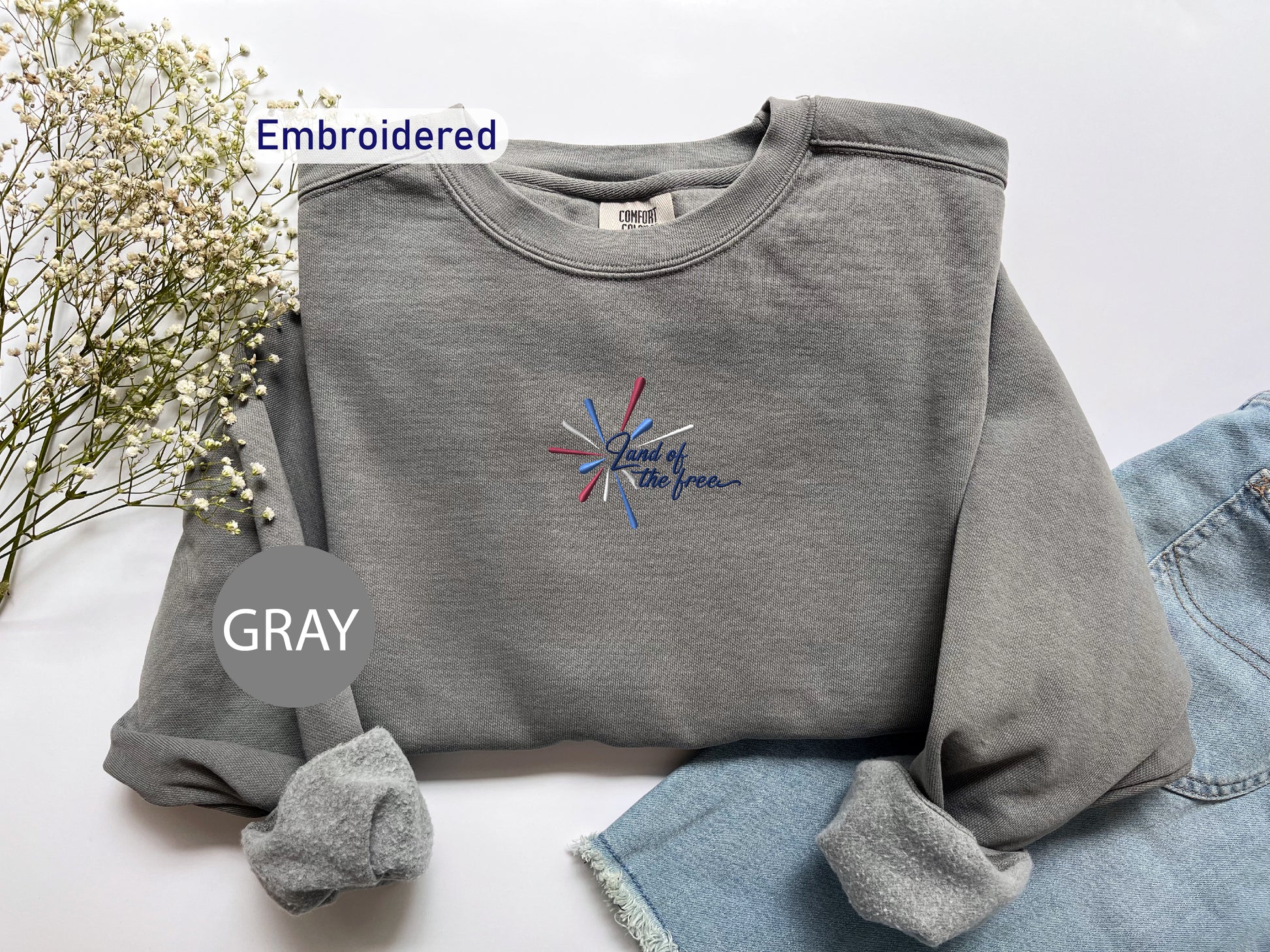 a gray sweatshirt with a patriotic design, a pair of denim jeans, and some flowers, all laid out on a white background.