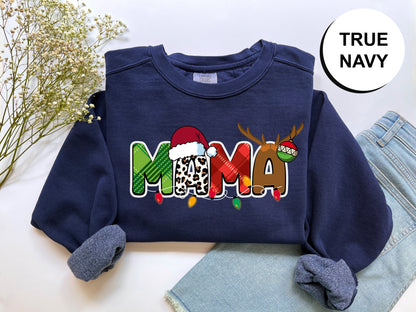 A navy blue sweatshirt with a colorful graphic design that says "MAMA" in a playful font, along with a Santa hat and reindeer. The sweatshirt is placed on a white surface, and there are some jeans visible in the background.