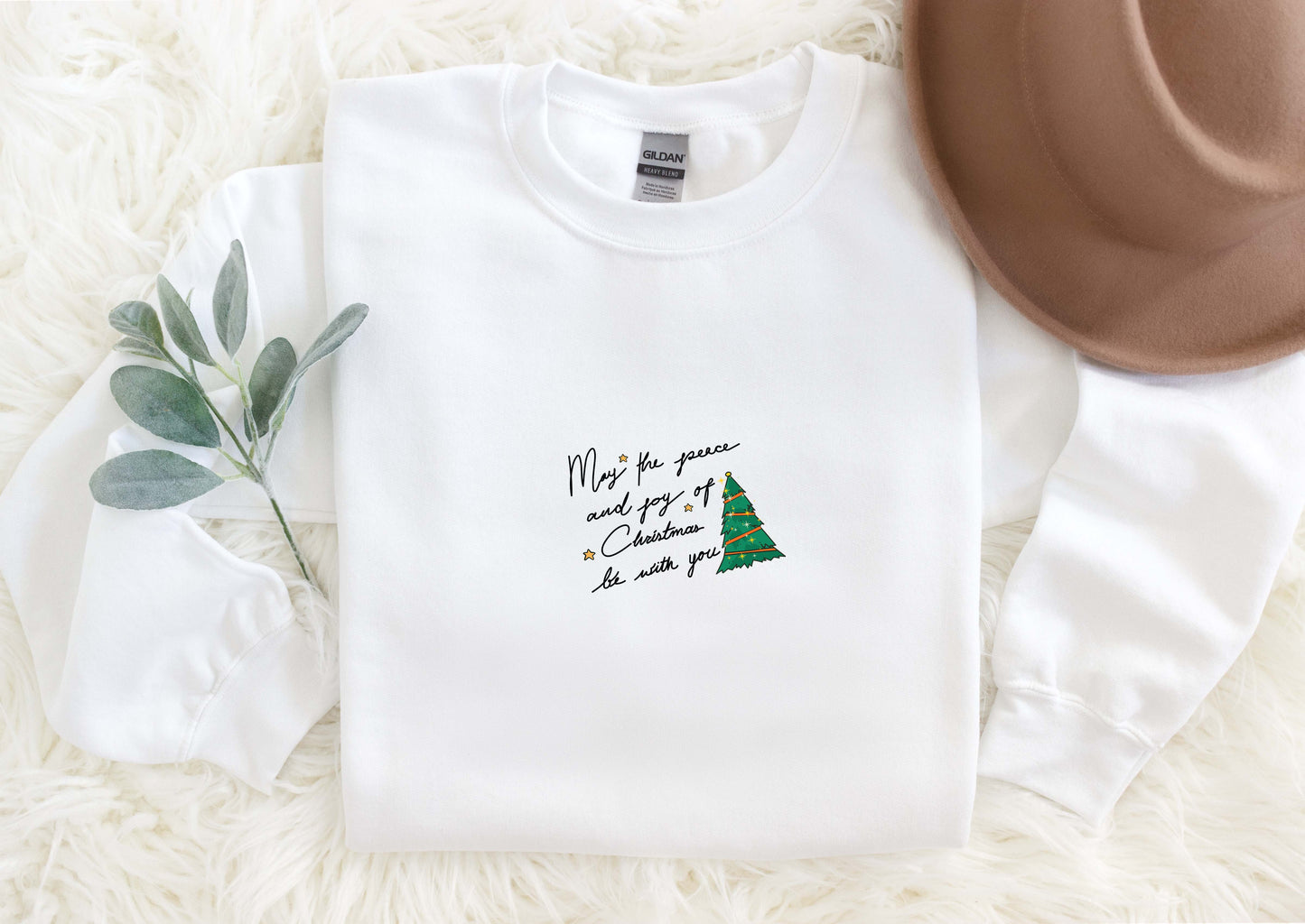 A white long-sleeved shirt with a Christmas tree design and a handwritten message, along with a brown hat, a green plant, and a white furry surface.