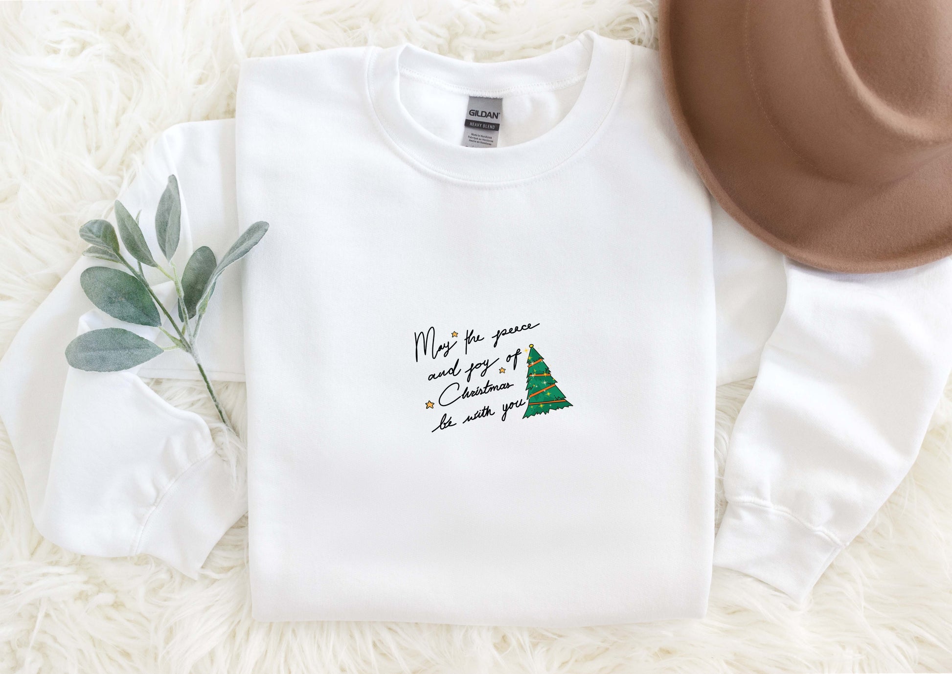 A white long-sleeved shirt with a Christmas tree design and a handwritten message, along with a brown hat, a green plant, and a white furry surface.