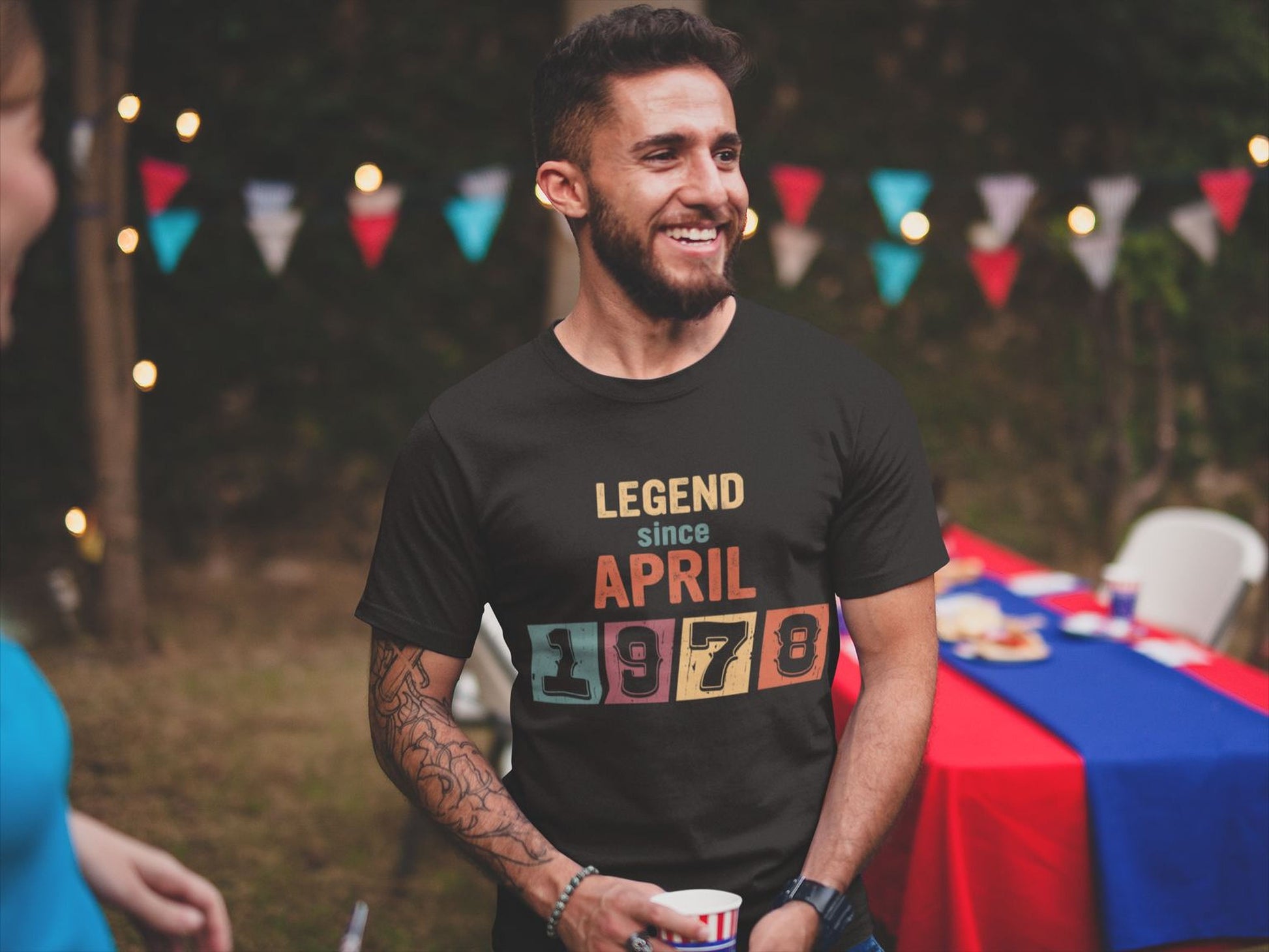 A man wearing a black t-shirt with the text "LEGEND since APRIL 1978" is smiling and standing in front of a table with a blue tablecloth.