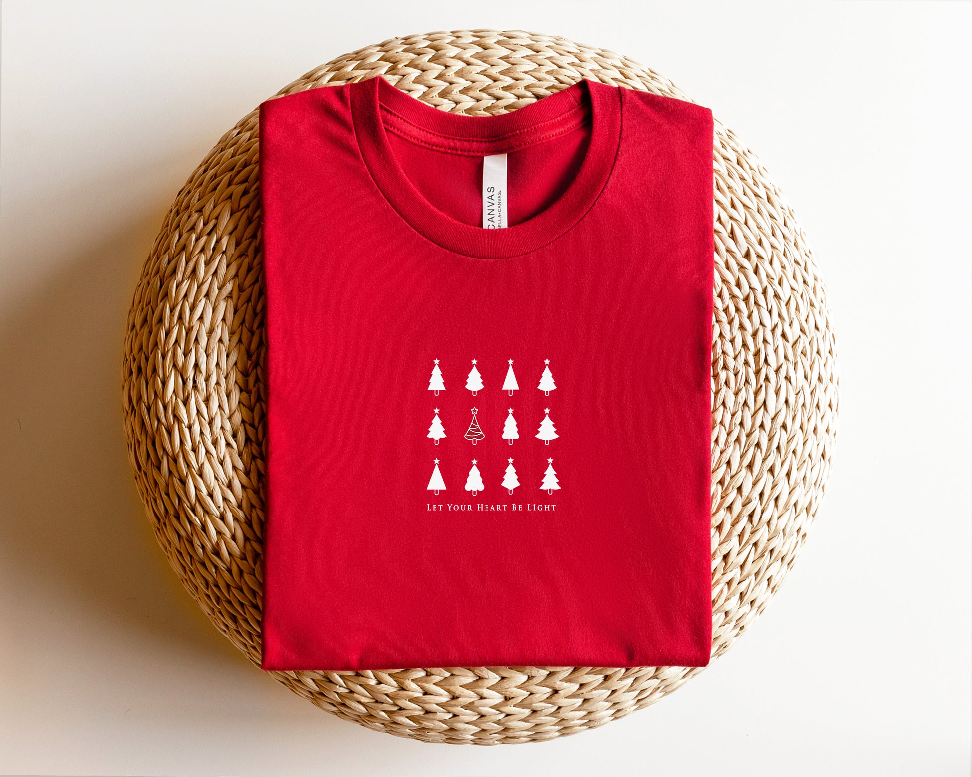 A red t-shirt with a white graphic design of Christmas trees and the text "LET YOUR HEART BE LIGHT" printed on it, placed on a woven basket.