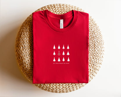 A red t-shirt with a white graphic design of Christmas trees and the text "LET YOUR HEART BE LIGHT" printed on it, placed on a woven basket.