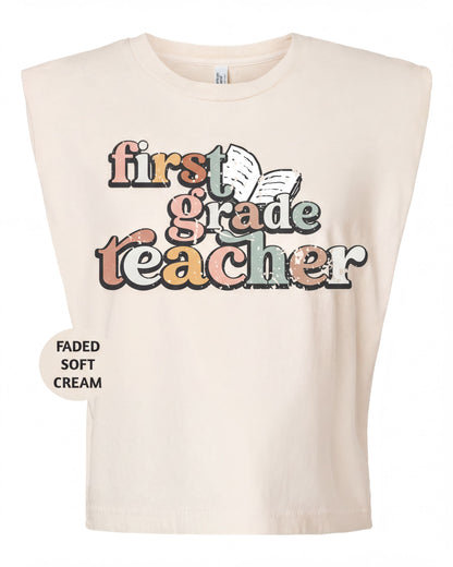 A beige t-shirt with a colorful graphic design that says "first grade teacher" in a playful font.