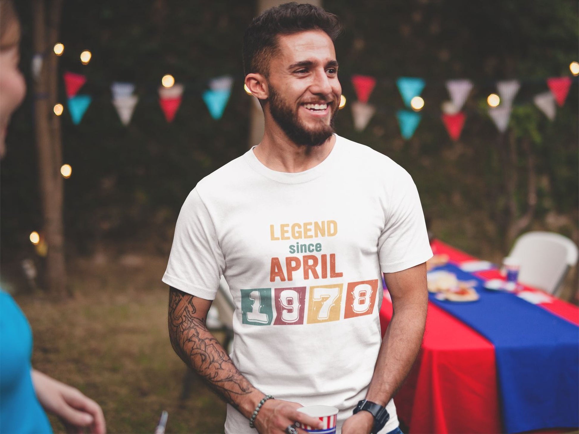 A man wearing a white t-shirt with the text "LEGEND since APRIL 1978" is smiling and standing in front of a table with a blue tablecloth.