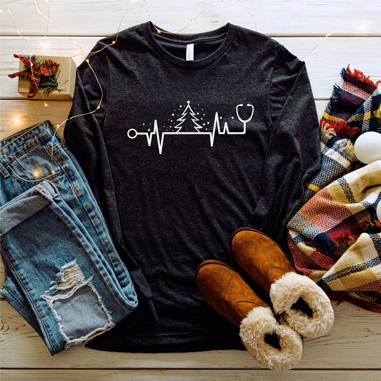 A casual outfit consisting of a black long-sleeved shirt with a white line design, a pair of distressed blue jeans, brown boots, and a plaid scarf is laid out on a wooden surface, along with some Christmas lights and a small gift box.