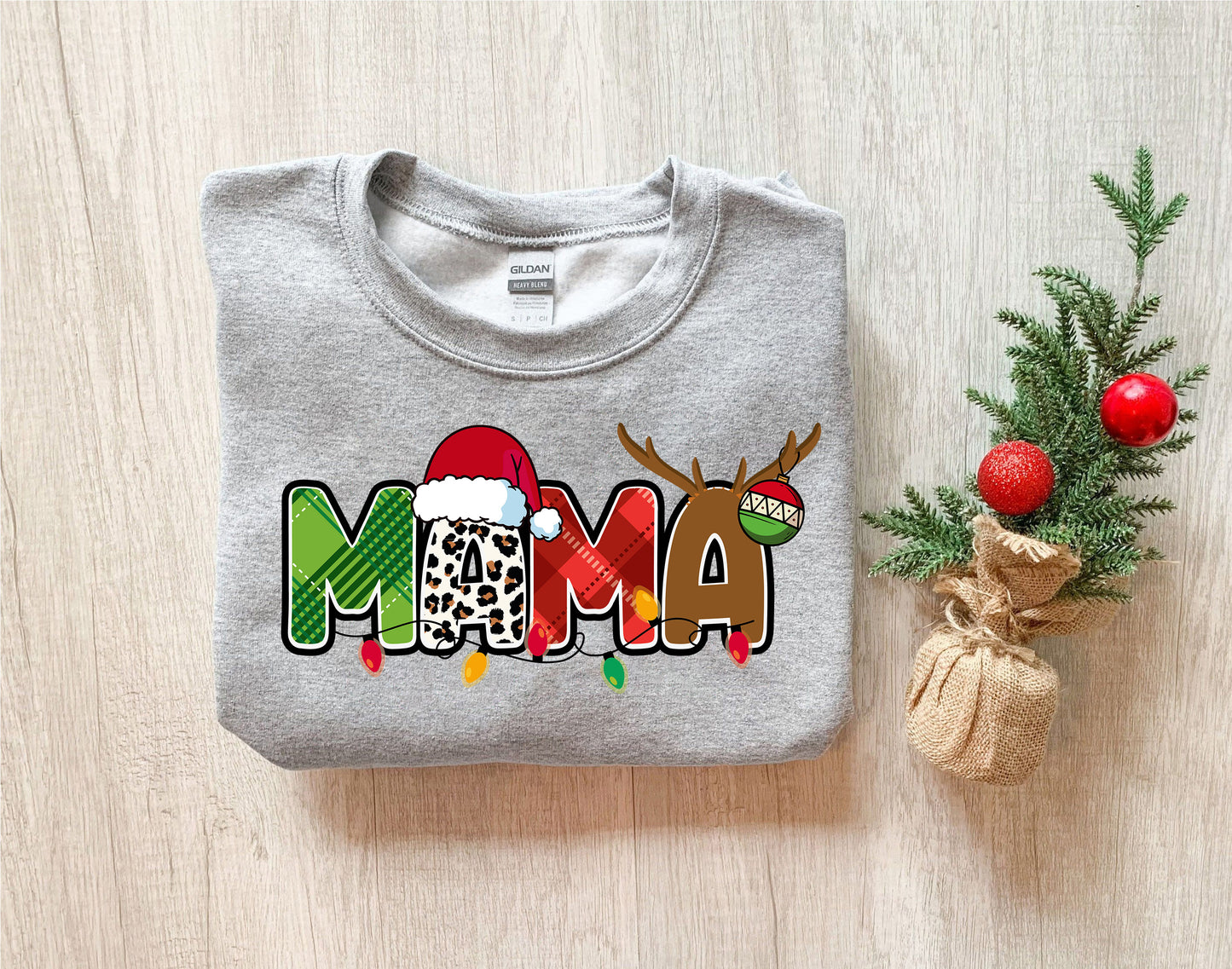 A gray sweatshirt with a Christmas-themed design, including the word "MAMA" in a stylized font, along with a Santa hat and antlers. The sweatshirt is placed next to a small Christmas tree and a burlap sack.
