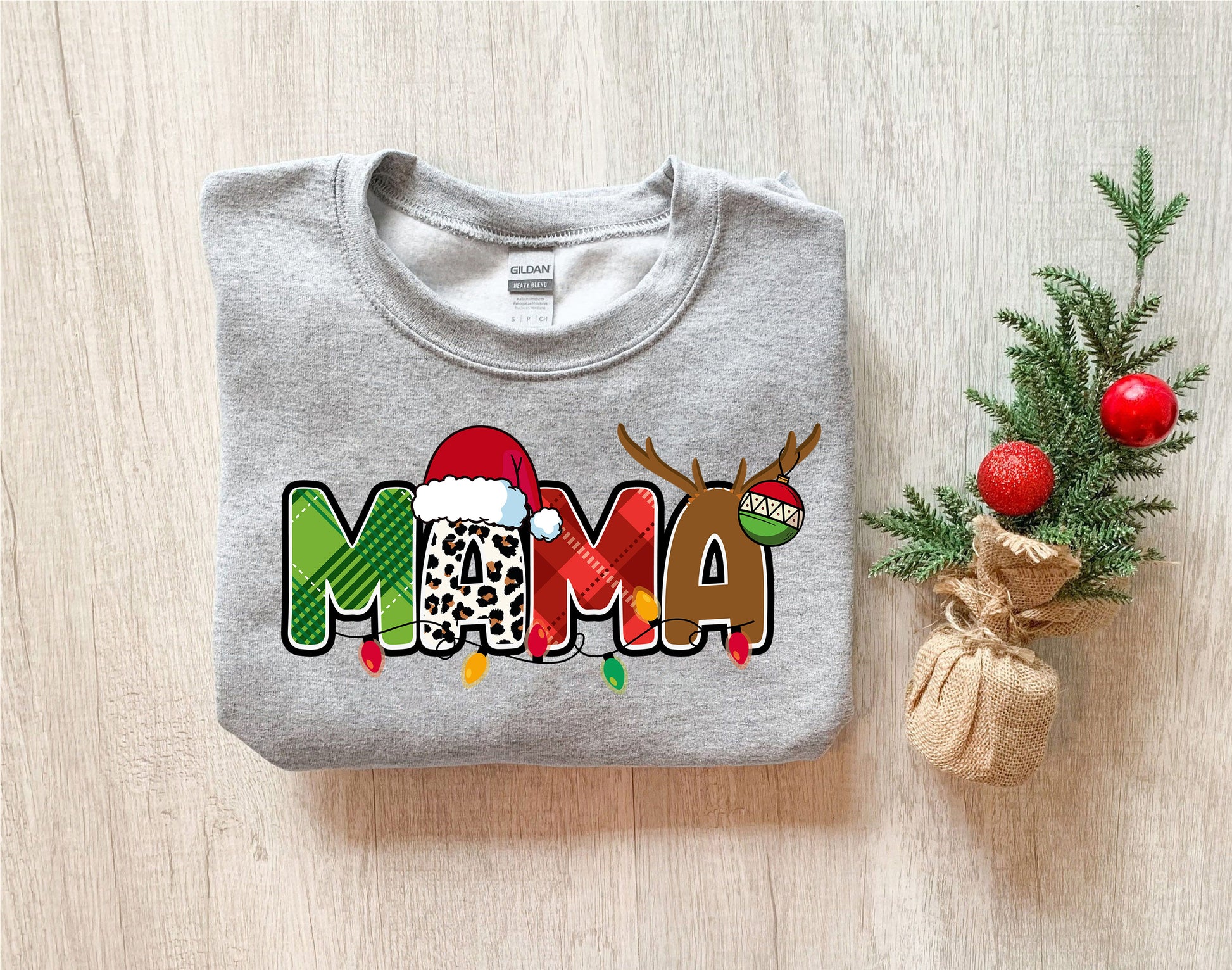A gray sweatshirt with a Christmas-themed design, including the word "MAMA" in a stylized font, along with a Santa hat and antlers. The sweatshirt is placed next to a small Christmas tree and a burlap sack.