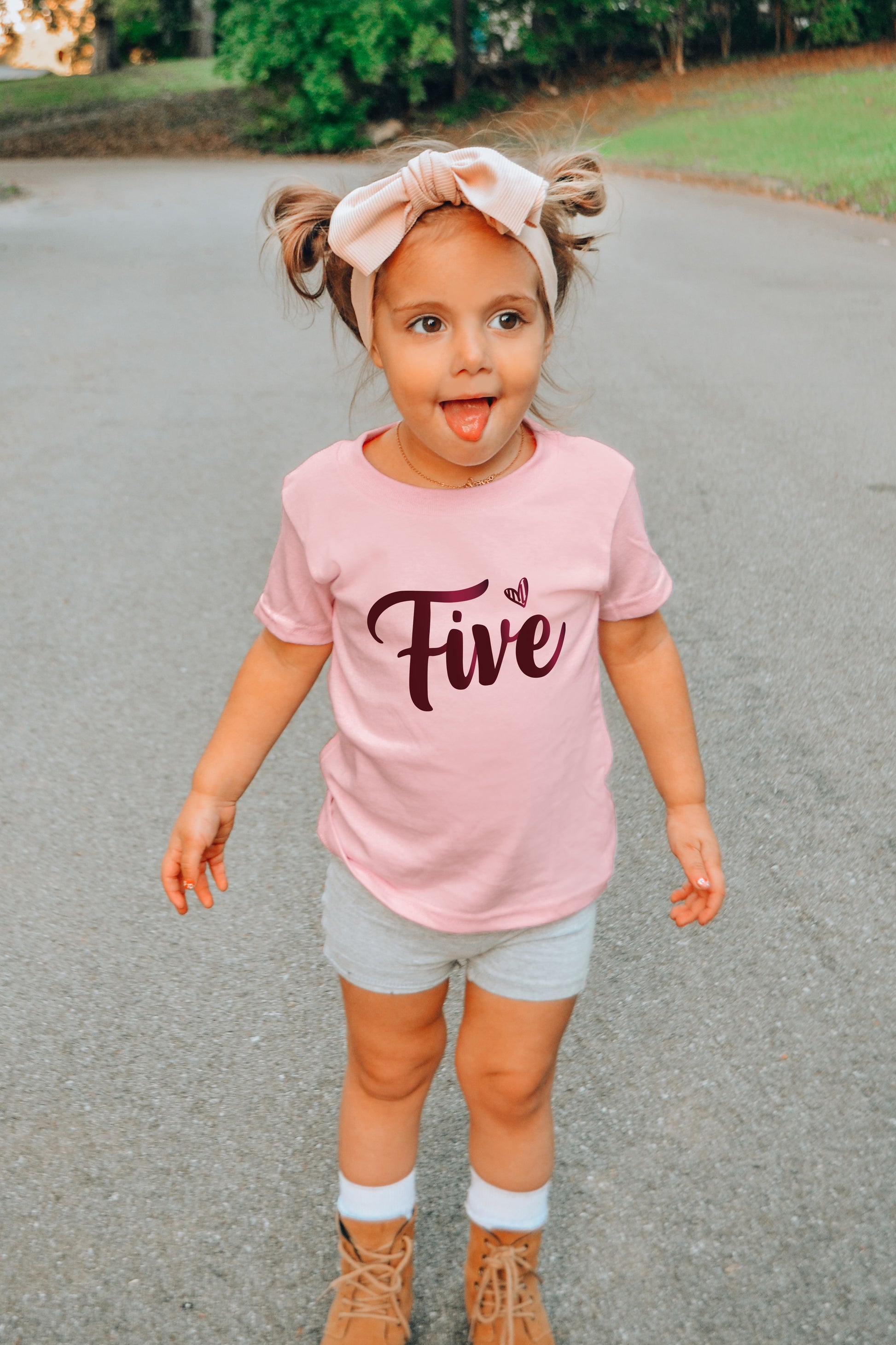 A young girl wearing a pink t-shirt with the number "Five" and a headband is standing on a paved surface, with her arms outstretched and her tongue sticking out.
