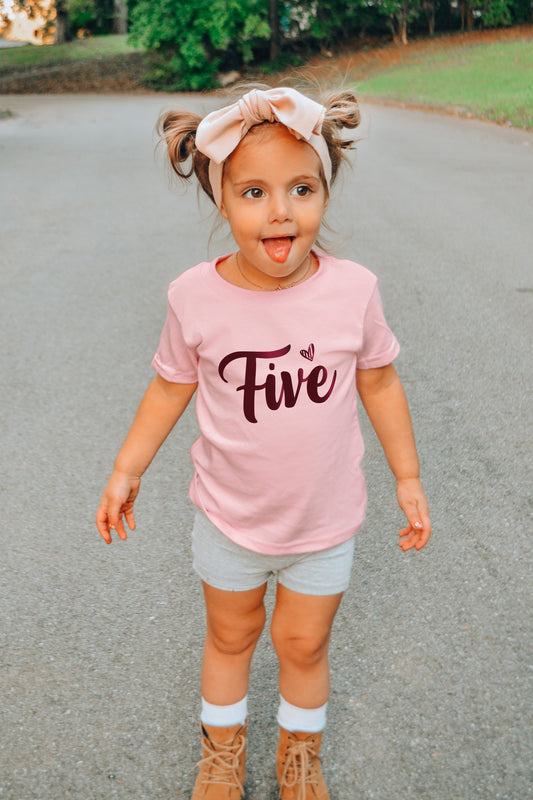 A young girl wearing a pink t-shirt with the number "Five" and a headband is standing on a paved surface, with her arms outstretched and her tongue sticking out.