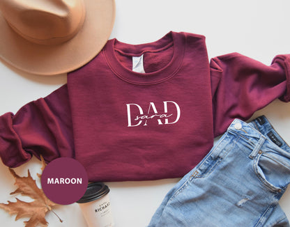 a casual outfit consisting of a burgundy sweatshirt with the text "DAD" printed on it, a pair of blue jeans, a brown hat, and a coffee cup.
