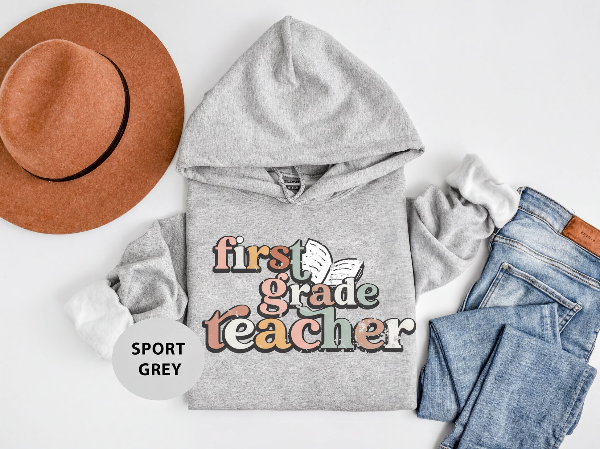 A gray hoodie with the text "first grade teacher" printed on it, along with a brown hat and a pair of blue jeans, all laid out on a white background.