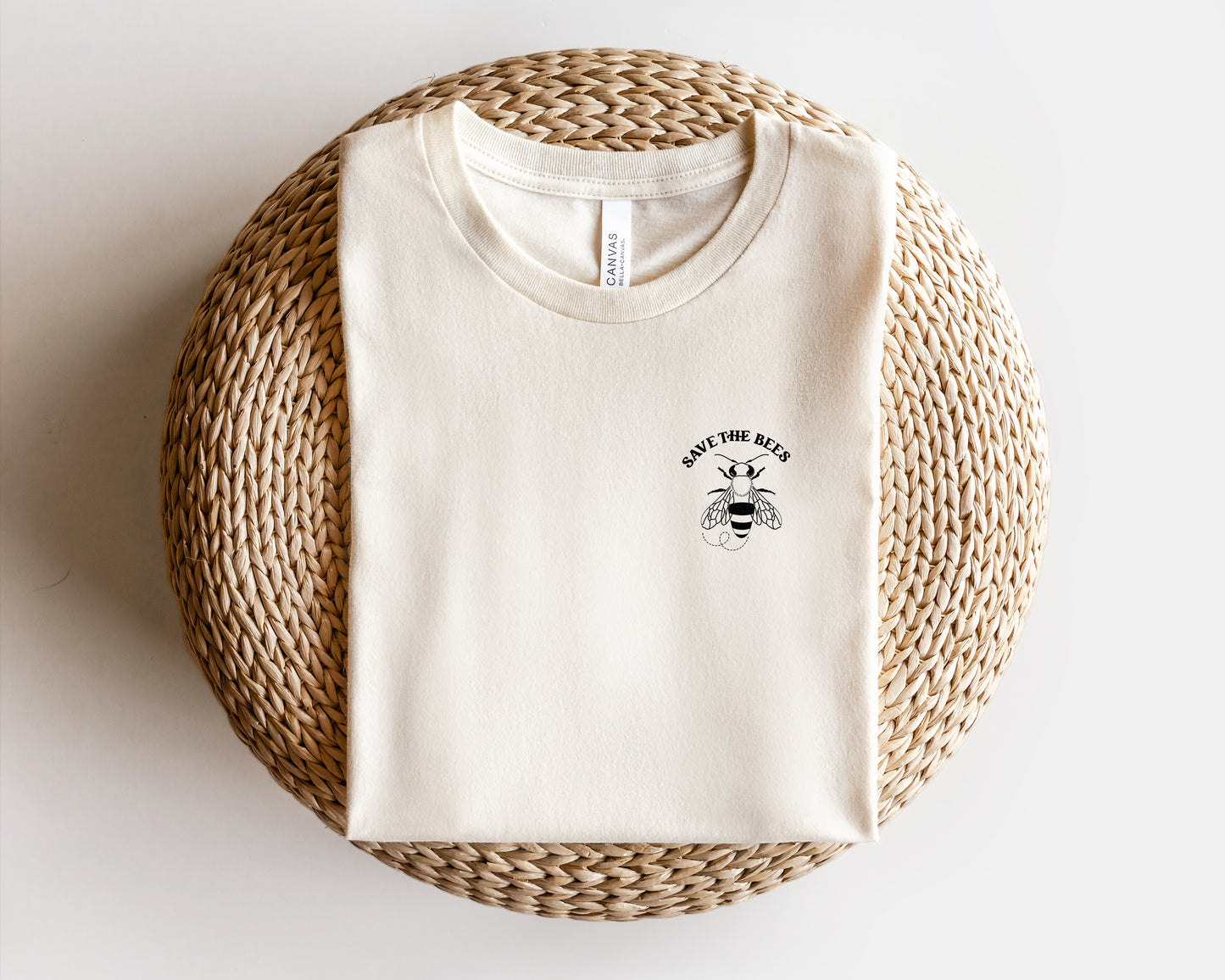 A white t-shirt with a graphic of a bee and the text "SAVE THE BEES" printed on it, laid out on a woven basket.