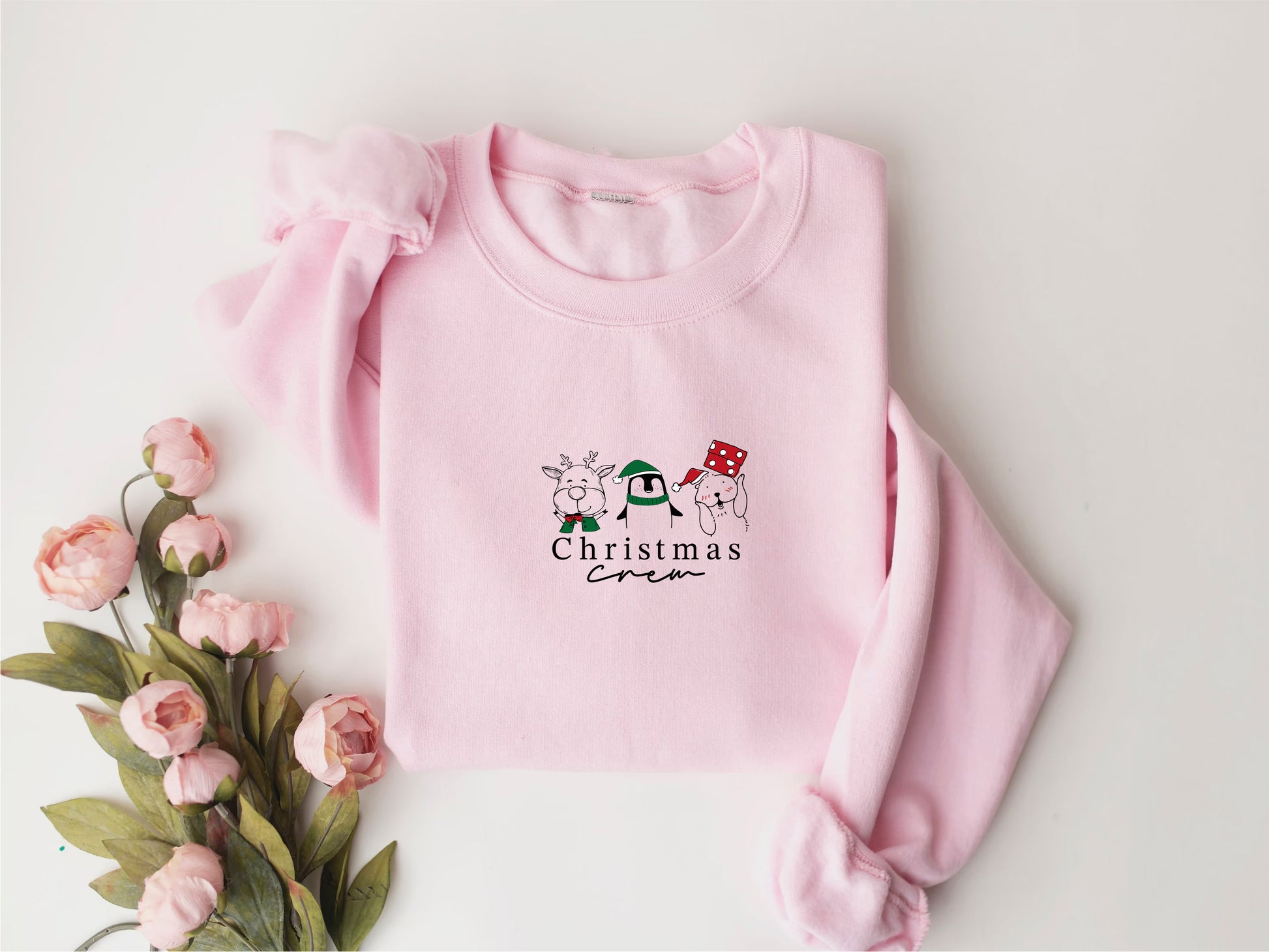 A pink crew neck sweater with a Christmas-themed design, including a reindeer, snowman, and a red and white striped scarf, is displayed alongside a bouquet of pink flowers.