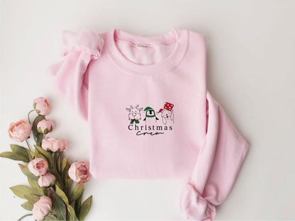 A pink crew neck sweater with a Christmas-themed design, including a reindeer, snowman, and a red and white striped scarf, is displayed alongside a bouquet of pink flowers.