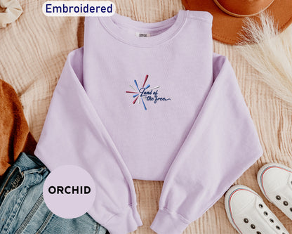 a purple sweatshirt with a patriotic design, a pair of jeans, a brown hat, and a pair of white sneakers, all laid out on a surface.