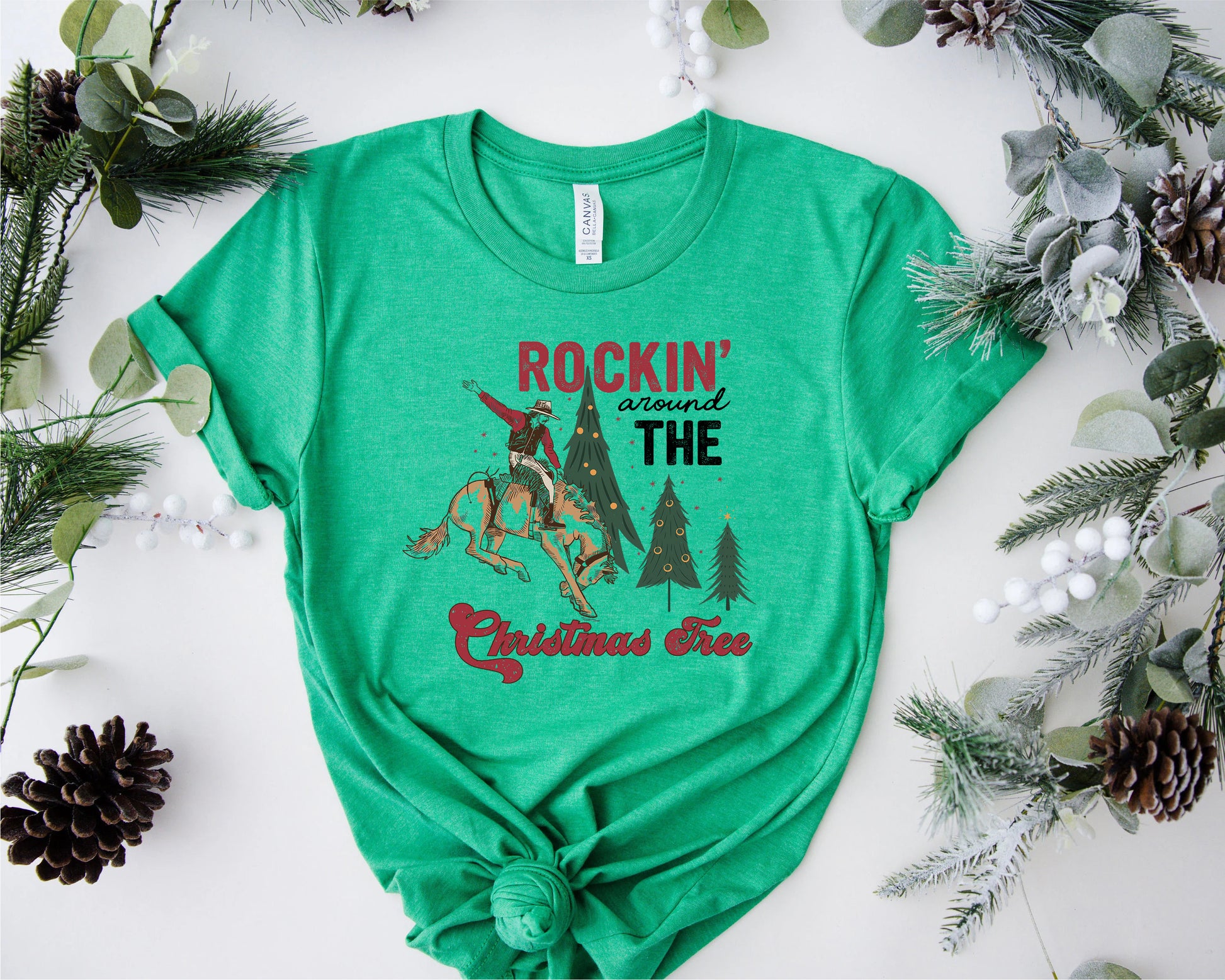 A green t-shirt with a graphic design featuring a cowboy on a horse, surrounded by Christmas trees and pine cones, with the text "ROCKIN' AROUND THE CHRISTMAS Tree" printed on it.