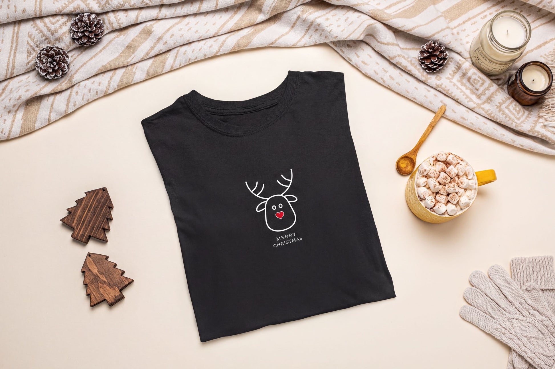 A black t-shirt with a white reindeer graphic and the text "Merry Christmas" is displayed on a table, accompanied by a mug of hot chocolate, pine cones, and a pair of gloves.