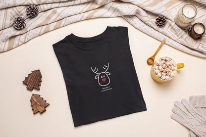 A black t-shirt with a white reindeer graphic and the text "Merry Christmas" is displayed on a table, accompanied by a mug of hot chocolate, pine cones, and a pair of gloves.
