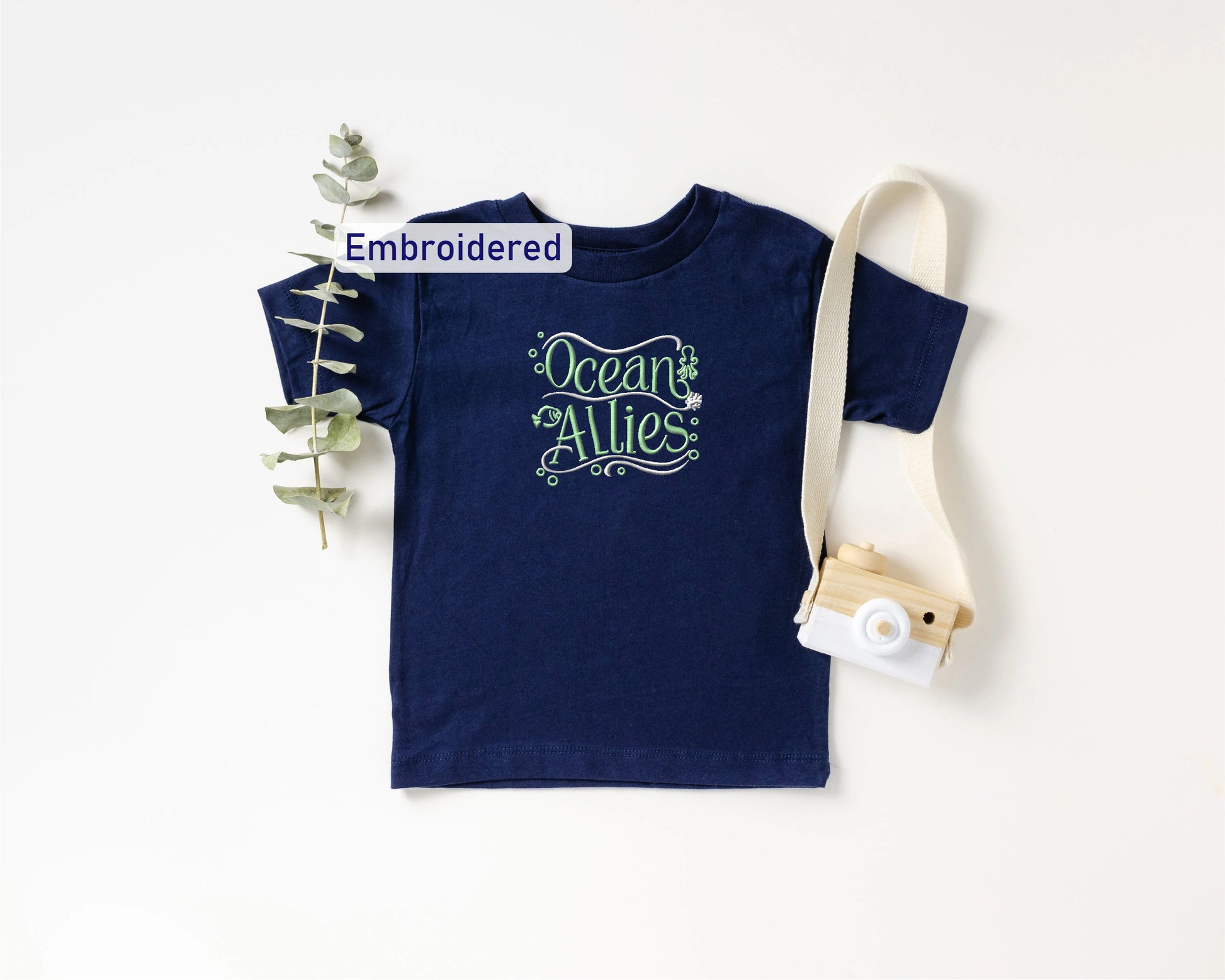 a navy blue t-shirt with the text "Ocean Allies" embroidered on it, along with a small camera and a plant.