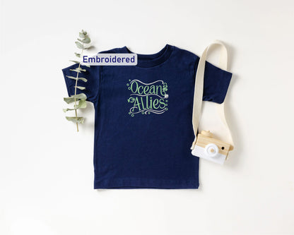 a navy blue t-shirt with the text "Ocean Allies" embroidered on it, along with a small camera and a plant.