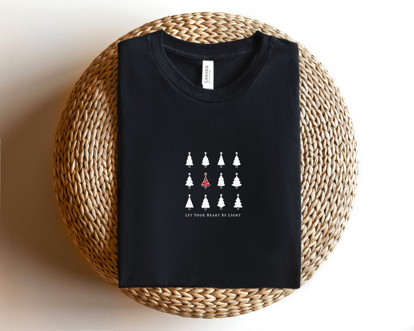 A black t-shirt with a white graphic design of Christmas trees and the text "Let your heart be light" printed on it, placed on a woven basket.