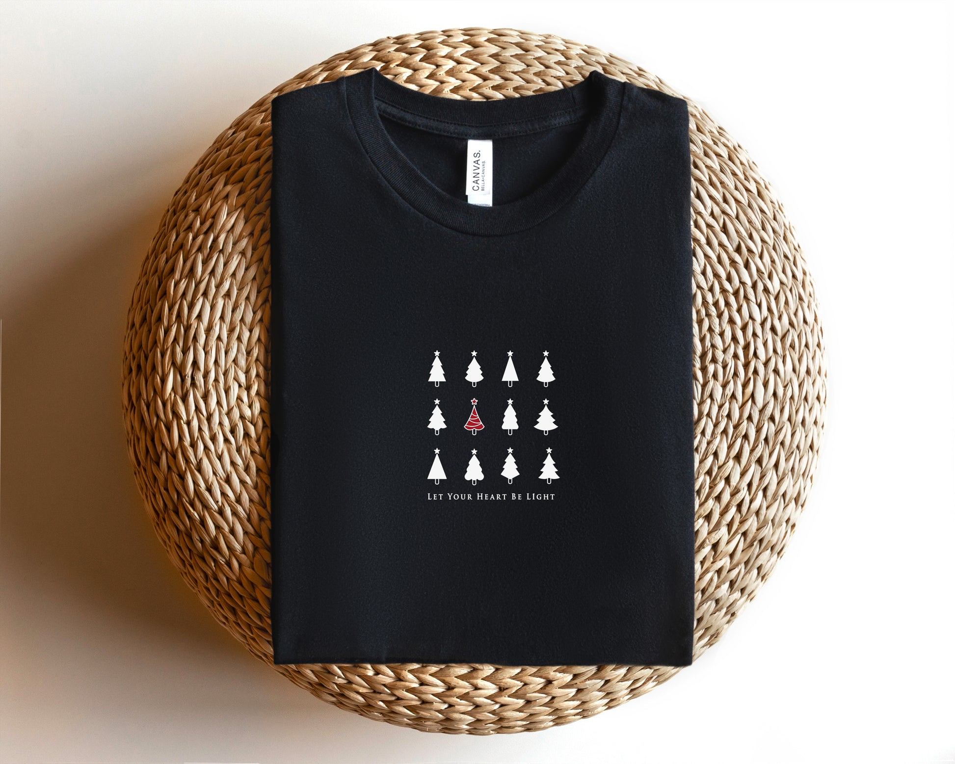 A black t-shirt with a white graphic design of Christmas trees and the text "Let your heart be light" printed on it, placed on a woven basket.