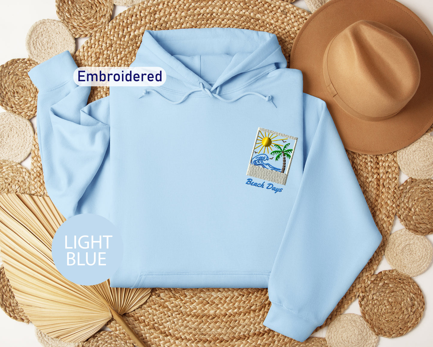 a light blue hoodie with a beach-themed patch on the front, a brown hat, and a straw hat, all arranged on a woven mat.