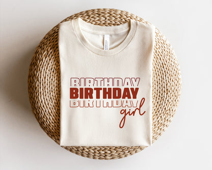 A white t-shirt with the text "BIRTHDAY BIRTHDAY girl" printed on it, laid out on a woven basket.