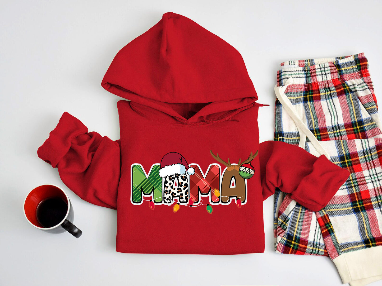 A red hoodie with the word "MAMA" printed on it, along with a pair of plaid pajama pants and a mug, all laid out on a white surface.