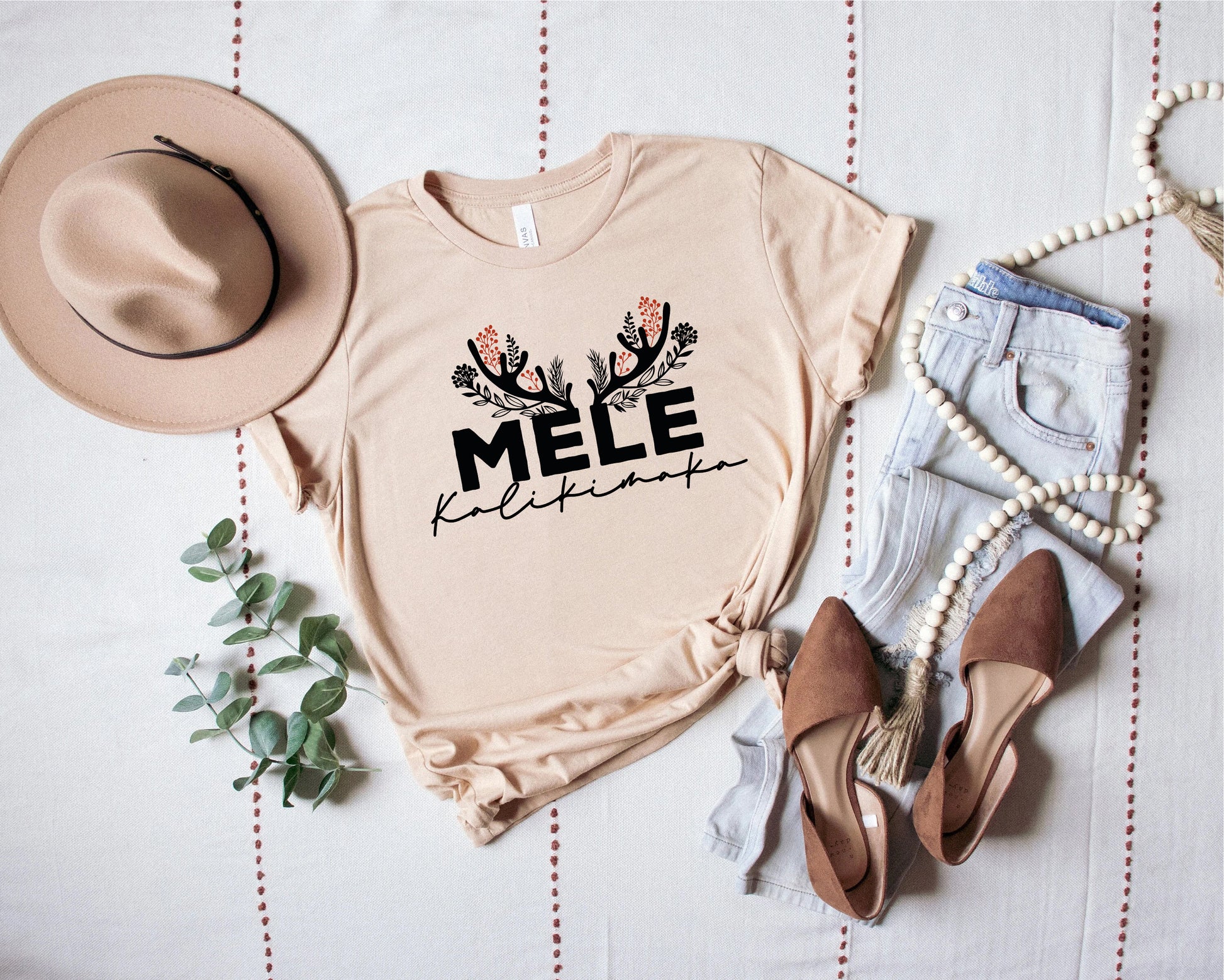 a casual outfit consisting of a beige t-shirt with a graphic design, a pair of jeans, a beaded necklace, and a hat.