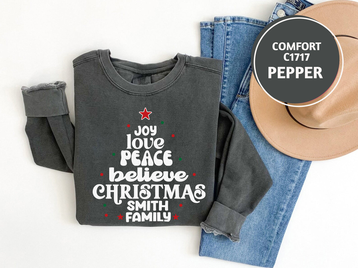 A gray sweatshirt with a Christmas tree design and text, a pair of blue jeans, and a beige hat are laid out on a white background.