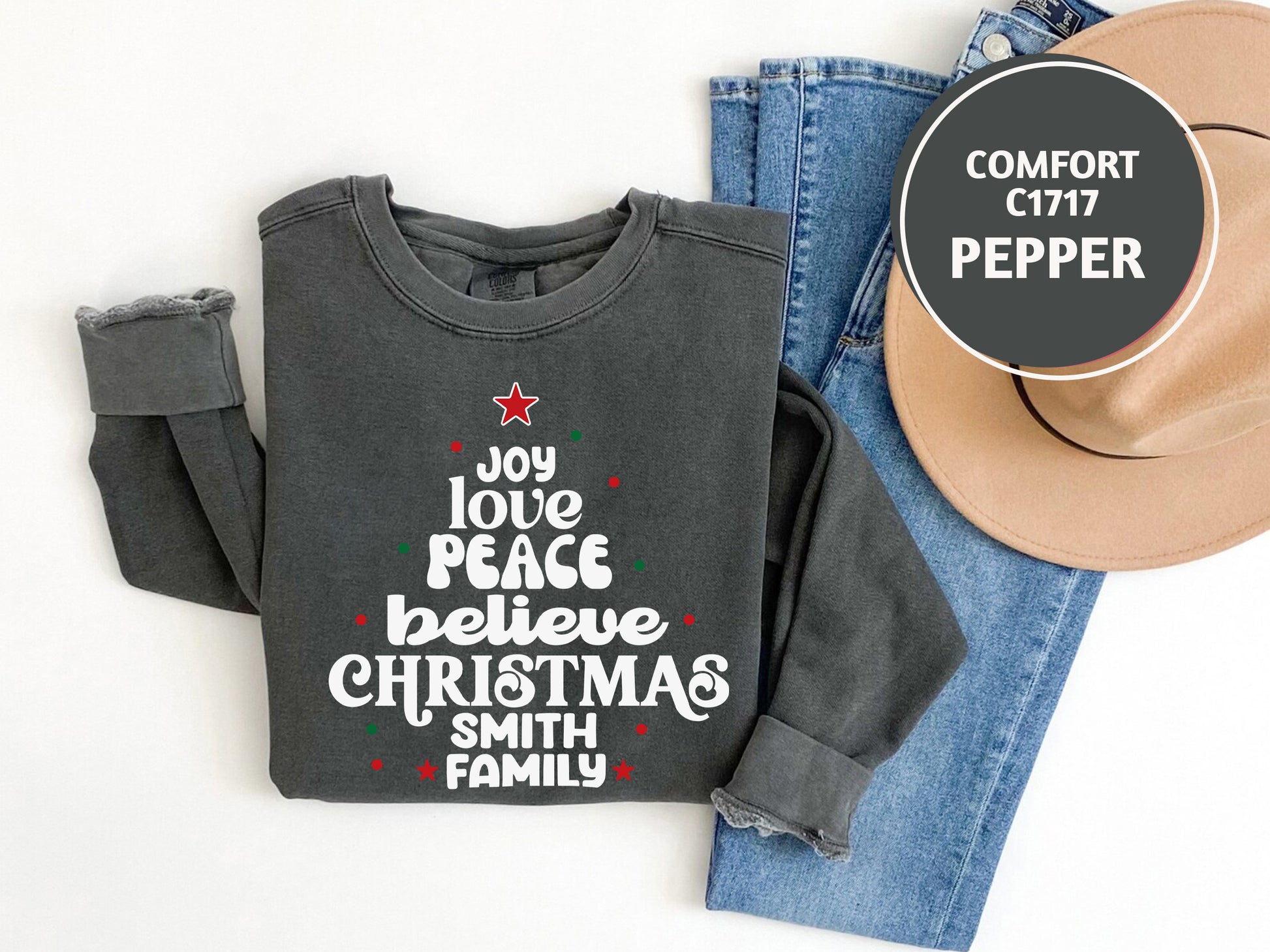 A gray sweatshirt with a Christmas tree design and text, a pair of blue jeans, and a beige hat are laid out on a white background.