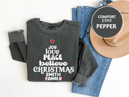A gray sweatshirt with a Christmas tree design and text, a pair of blue jeans, and a beige hat are laid out on a white background.