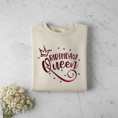 a white sweatshirt with a red and purple design that says "Birthday Queen" and stars. The sweatshirt is laid out on a marble surface, and there is a bouquet of white flowers next to it.