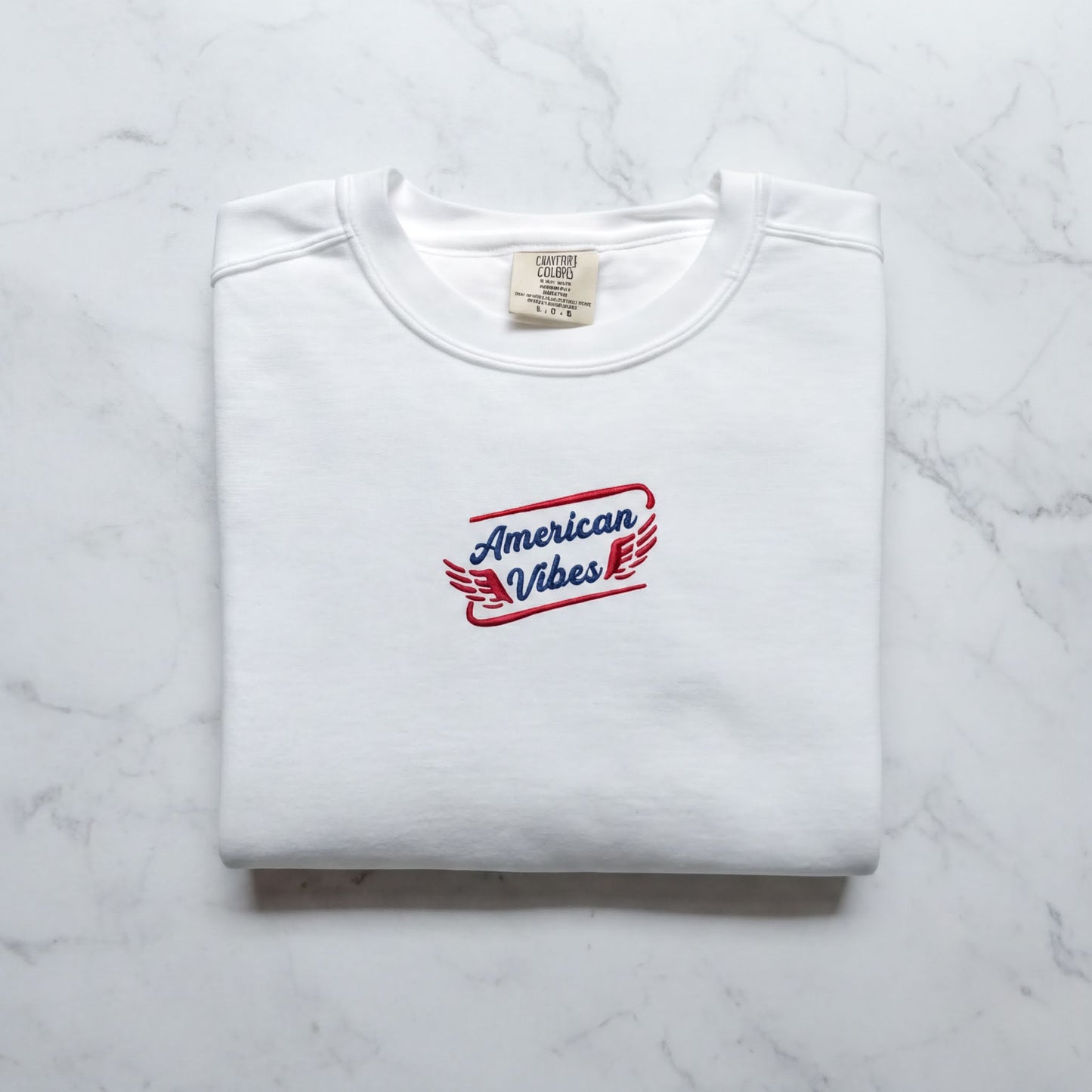 A white t-shirt with a red and blue logo that says "American Vibes" on it, laid out on a marble surface.