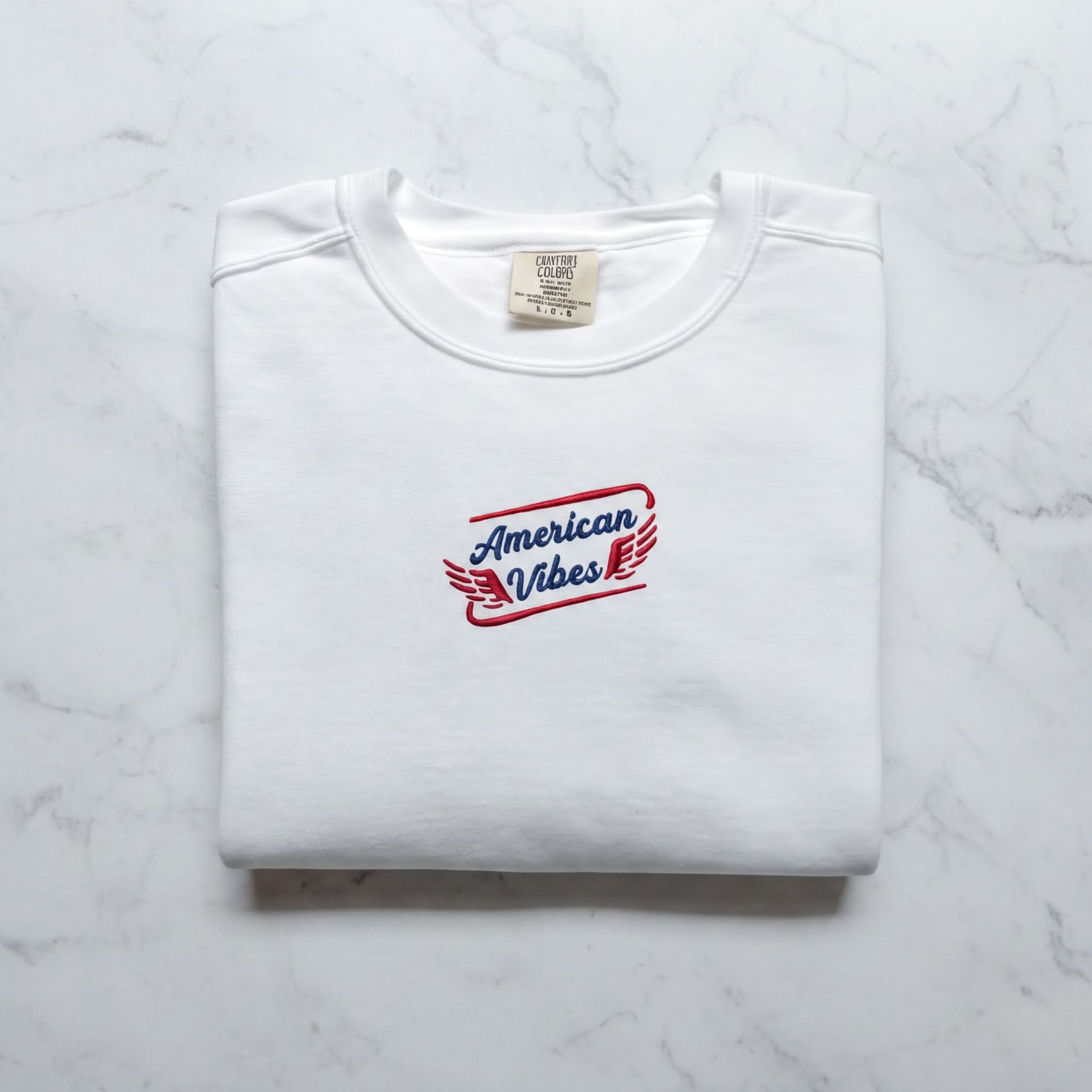 A white t-shirt with a red and blue logo that says "American Vibes" on it, laid out on a marble surface.