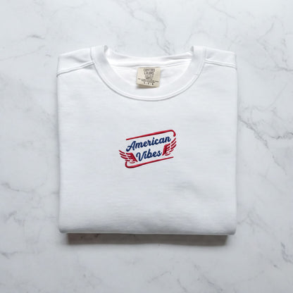 A white t-shirt with a red and blue logo that says "American Vibes" on it, laid out on a marble surface.