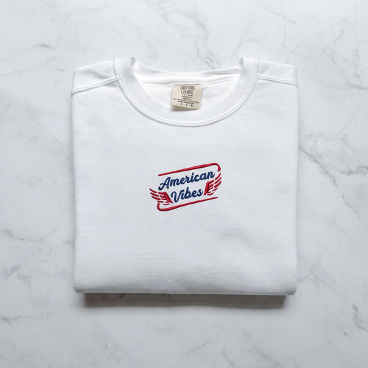 A white t-shirt with a red and blue logo that says "American Vibes" on it, laid out on a marble surface.