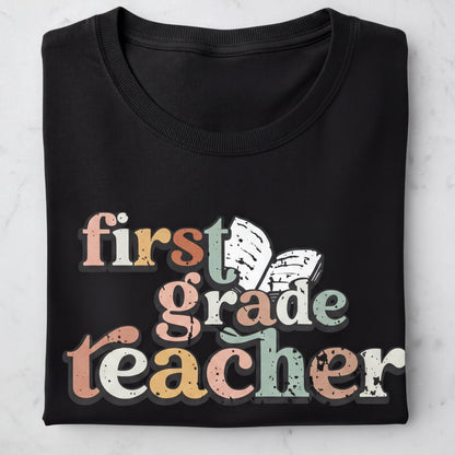 A black t-shirt with the text "first grade teacher" printed on it in a colorful, distressed font.