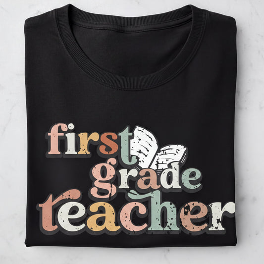 A black t-shirt with the text "first grade teacher" printed on it in a colorful, distressed font.