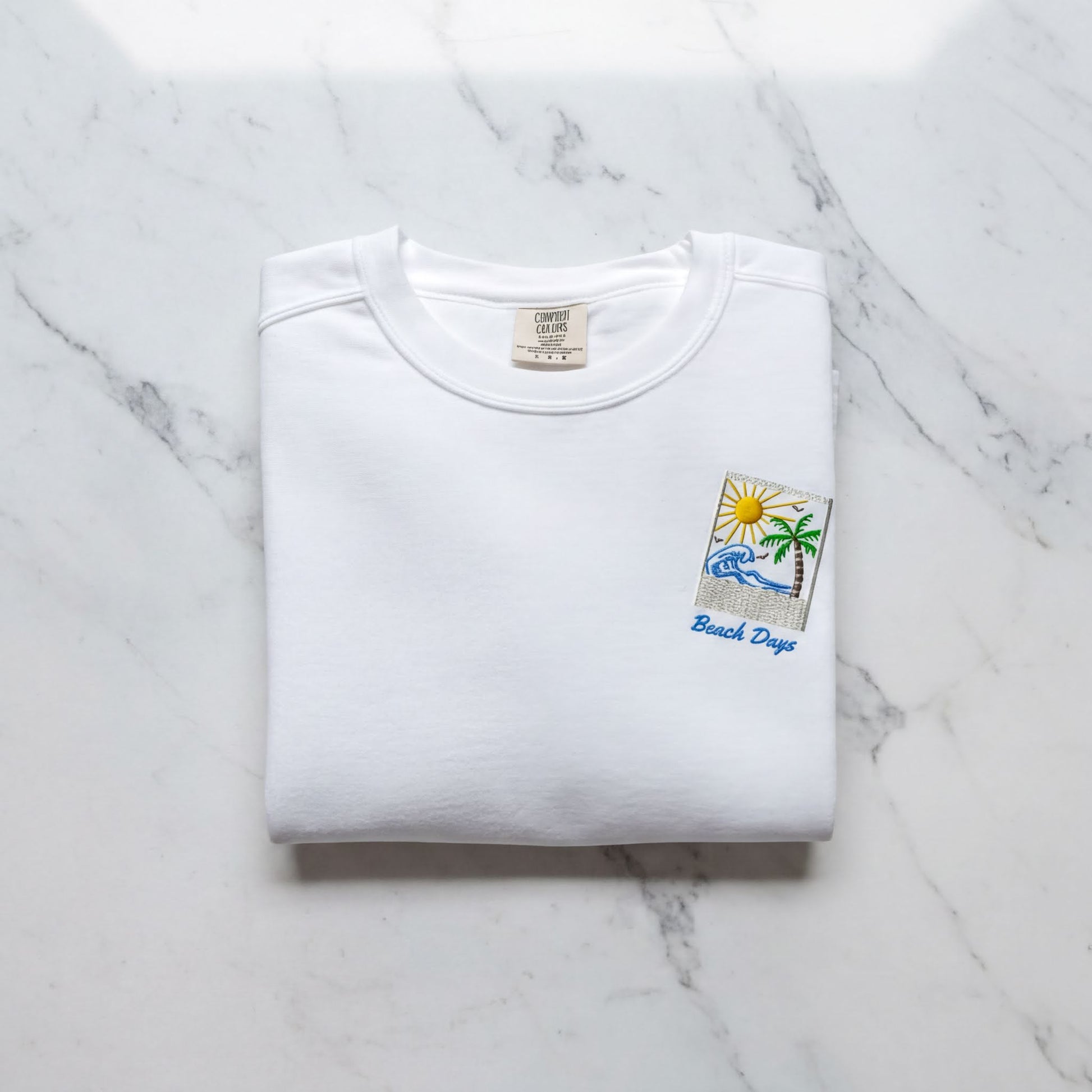 A white t-shirt with a beach-themed design and the text "Beach Days" printed on it, laid out on a marble surface.