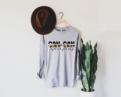 a gray long-sleeved t-shirt with the text "CAN CAN" printed on it, hanging on a wooden hanger. Next to the t-shirt is a brown hat, and there is a potted plant in the background.