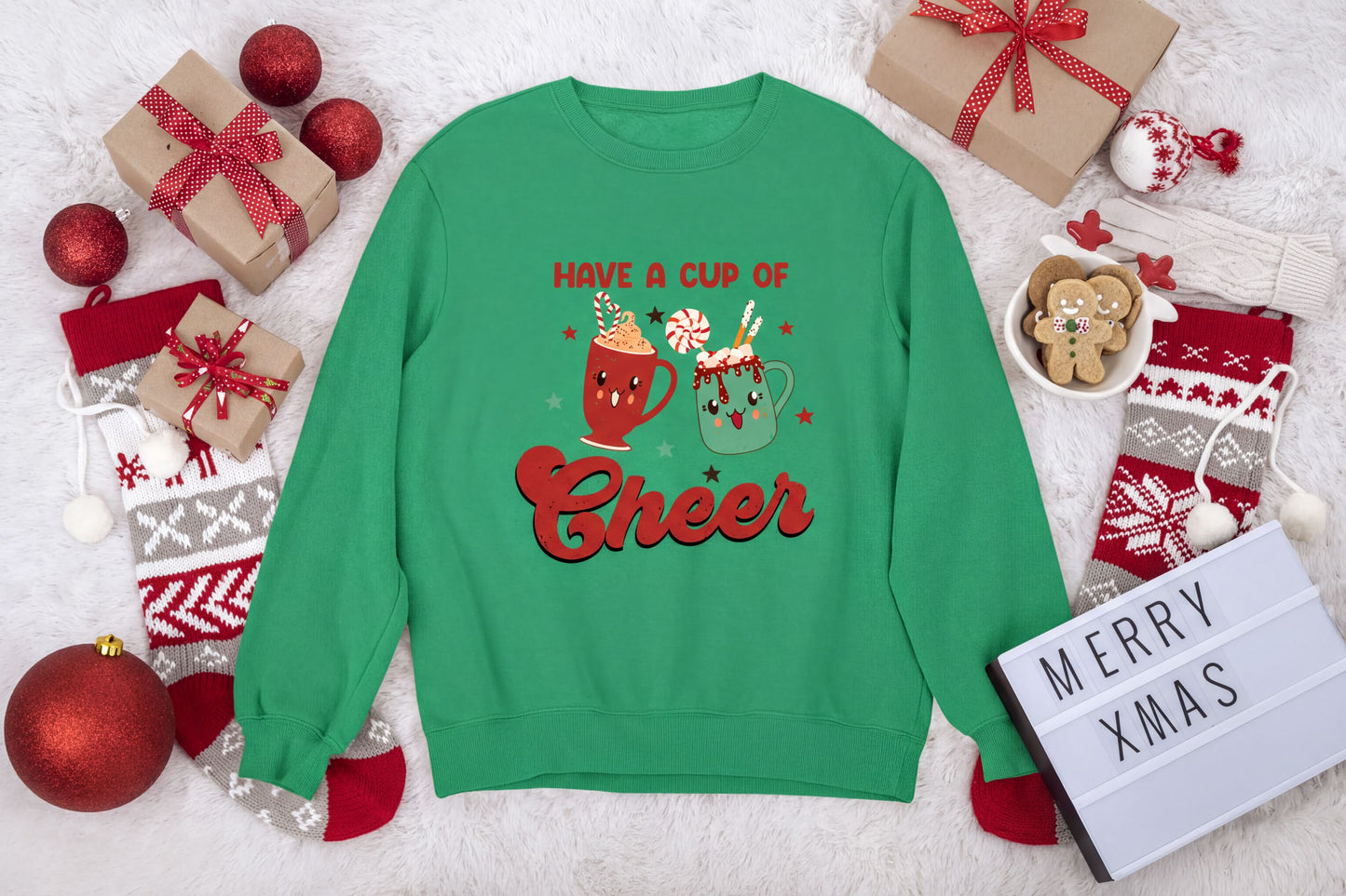 A green sweatshirt with a graphic design of two mugs and the text "Have a cup of cheer" is surrounded by various Christmas-themed items, including gift boxes, ornaments, and a "Merry Xmas" sign.
