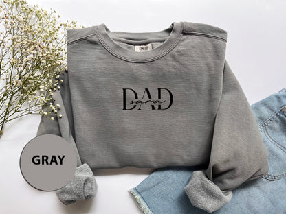 A gray sweatshirt with the text "DAD" printed on it, along with a gray circle labeled "GRAY" and some flowers in the background.